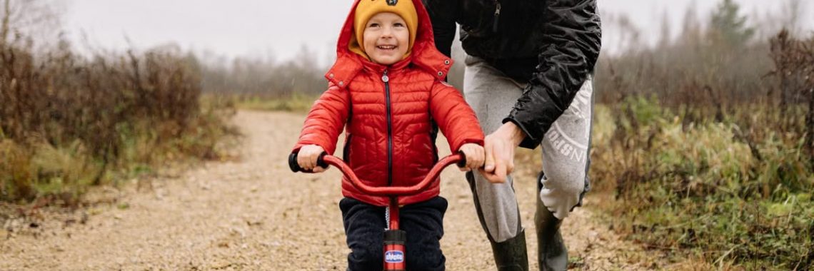 A father joyfully helps his child ride a bicycle on a gravel path in an autumn setting.